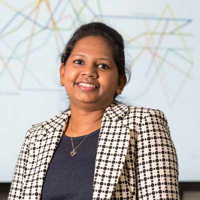 A woman with long dark hair sits on a bright green couch, smiling while using a laptop covered in various black and white stickers. She wears a black dress, and the background features a modern, minimalist office setting.