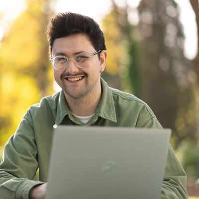 A woman with long blonde hair sits in an orange chair, smiling while speaking to another person whose back is facing the camera. She wears a light sweater, and theres a laptop and glass of water on a table in front of her.