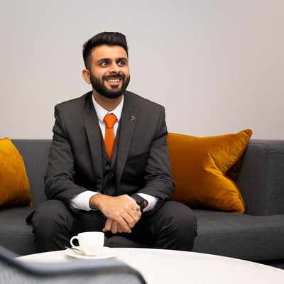 A man wearing a dark suit with an orange tie stands smiling in a modern office lobby with wooden floors and open double glass doors in the background. An orchid is visible on a table to the left.
