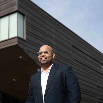 A person with glasses and a mustache, wearing a green jacket, is sitting outdoors at a table with a laptop. Theres a blue mug and a pen next to the laptop. Trees and foliage are visible in the blurred background.