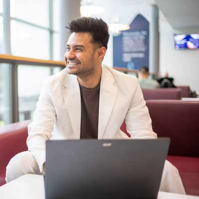 A person with long blonde hair is smiling while seated, wearing a light beige sweater. They are facing another person and have a laptop in front of them on a table. The background has a geometric pattern.
