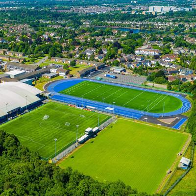 Aerial view of a sports complex featuring a large indoor sports facility, blue athletics track, multiple soccer fields, lush green trees in the foreground, and nearby residential buildings in the background.