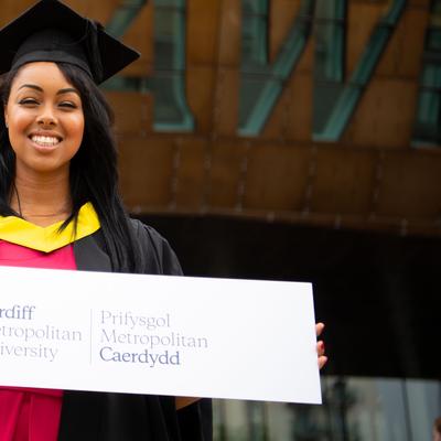 A Cardiff Met student, in graduation cap and gown, stands holding a sign featuring the Cardiff Met logo and name