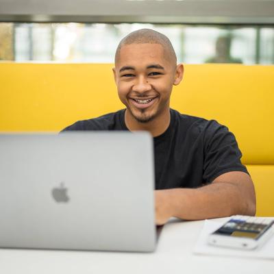 A person is smiling while sitting at a table with a laptop in front of them. They are in a bright, modern setting, with a yellow cushioned seating area behind them.