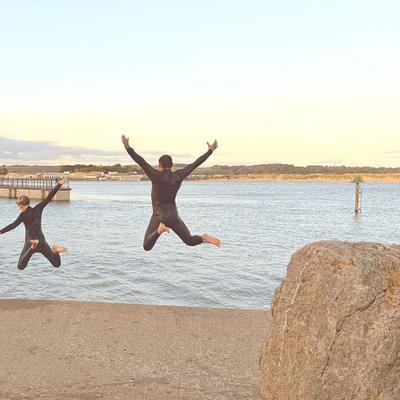 Two people wearing wetsuits jump into the water from a concrete dock