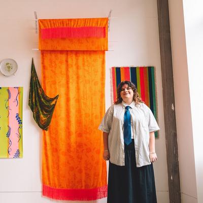 A young man in a shirt and tie stands beside a display of colourful textiles