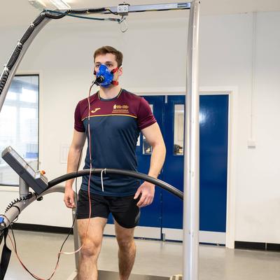 A man is walking on a treadmill wearing a blue mask for a fitness test. A woman stands nearby, operating a computer connected to the equipment. Both are in a lab setting wearing maroon and navy sports attire.