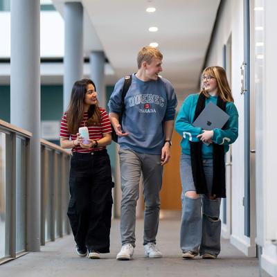 Three young people walk together on a balcony. One is holding a laptop computer.