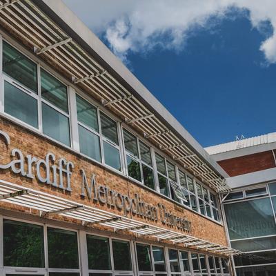 A building with large windows featuring the sign Cardiff Metropolitan University on a brick wall. A blue sky with clouds is visible above.