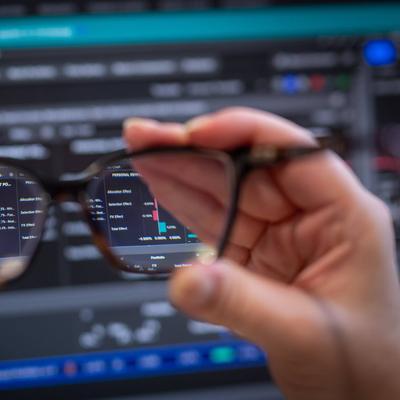 A computer screen displaying stock market data and graphs, viewed through a pair of glasses held in someone's hand.