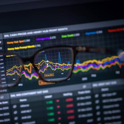A computer screen displaying stock market data and graphs, viewed through a pair of glasses held in someone's hand.