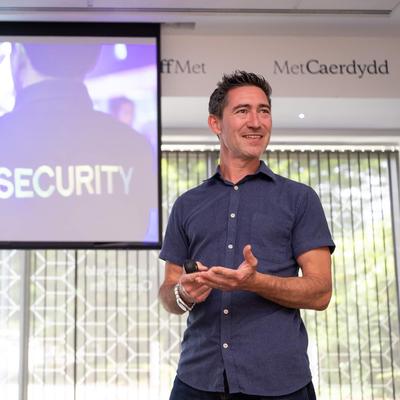 A man stands in front of a screen giving a presentation. The screen shows a person wearing a shirt with SECURITY printed on the back.