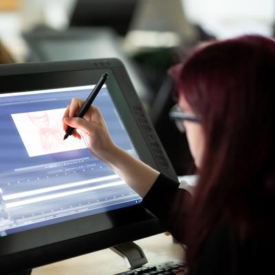 A person with long hair is using a stylus to draw on a large digital tablet. The screen displays graphic design software with artwork in progress. Other people are visible in the background, working at similar stations.