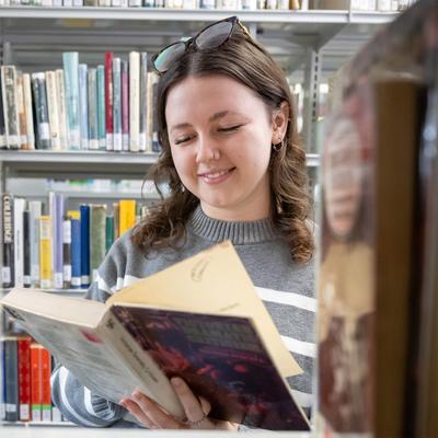 A woman in a striped sweater is smiling while reading a book in a library. She has sunglasses on her head and stands surrounded by shelves filled with books.