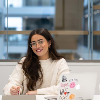 A woman with long hair and glasses sits at a desk smiling, using a stylus on a tablet. She is in front of a laptop with various stickers. Next to her, a mannequin displays a white T-shirt with colourful text. The setting is a modern office.
