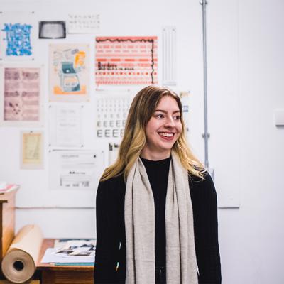 A person with long hair and a scarf stands smiling in an art studio. Behind them is a wall with various vintage posters and prints. A wooden chest of drawers and rolls of paper are visible in the foreground.