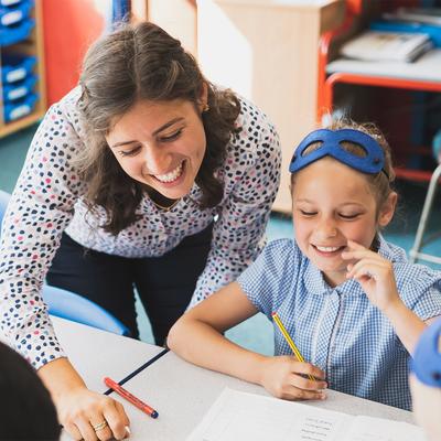 A teacher leans over a classroom table, smiling and engaging with two elementary-aged students in school uniforms. The students are seated and holding pencils, with colourful storage bins visible in the background.