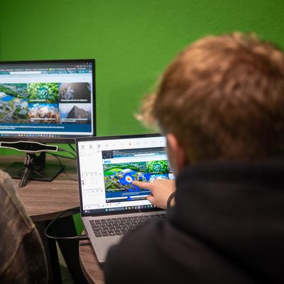 Two people are looking at computer screens in an office. One person points at a laptop displaying a design of a landscape, while a larger monitor shows similar images. The room has bright green walls.