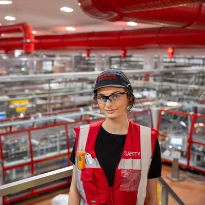 A person wearing safety gear and a cap stands in a large industrial facility with extensive machinery and red overhead pipes. The person is wearing a red vest labelled SAFETY and safety goggles, and looks toward the camera.