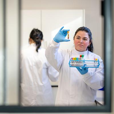 A female scientist in a white lab coat and blue gloves examines a test tube held up to the light. Another person in a lab coat works in the background. Various test tubes are organized in a rack. The scene is viewed through a glass window.