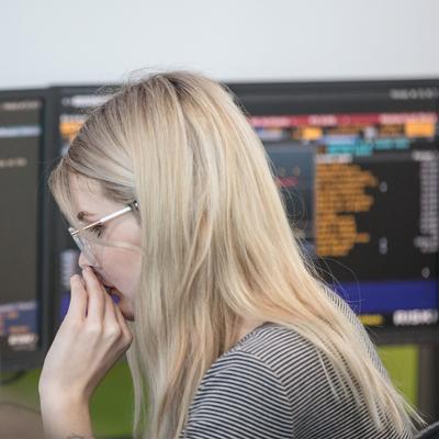 A woman with blonde hair and glasses is intently focused on monitors displaying complex data and graphs. She wears a striped shirt and appears to be in a busy office environment.