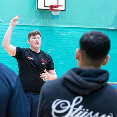 A person stands in a gym, gesturing with one hand raised, facing a group. The back of one person is visible, wearing a shirt labelled Coach. A basketball hoop is in the background. The setting is casual and suggests a team gathering or discussion.