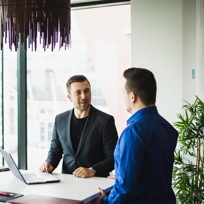 Two men are seated at a white table in a modern office with large windows. One is using a laptop, and they appear to be having a conversation. Natural light fills the room, and theres a potted plant nearby.