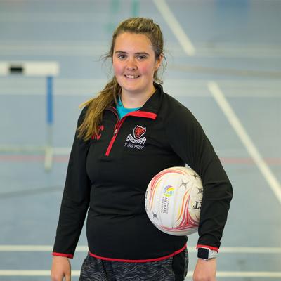 A person in sports attire stands in an indoor court, holding a netball with one hand. The court has visible markings and a hurdle in the background. The person is smiling and wearing a black jacket with a logo.