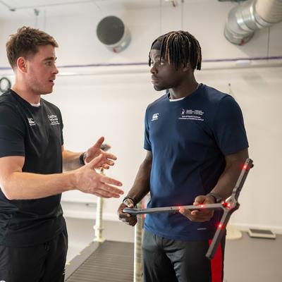 Two men are engaging in conversation in a lab setting. One is gesturing with his hands, while the other holds a device with sensors. Both are wearing athletic gear. A treadmill and office equipment are visible in the background.