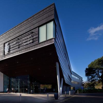 Modern building with angular architecture, featuring a large sign that reads Prifysgol Metropolitan Caerdydd. The structure has a mix of dark panels and white walls, with glass windows. Trees and a blue sky are visible in the background.