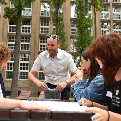 Four people sit at a wooden table outdoors. A man wearing a name badge labelled Gary stands next to them.