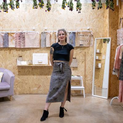 A woman stands smiling in a cozy, wood panelled boutique. She wears a black top and a long skirt. The room features dried flowers, a chair with a patterned pillow, art on the walls, and display shelves with items.