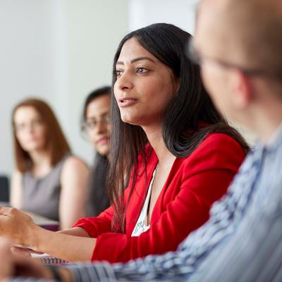 A woman in a red blazer speaks during a meeting, surrounded by colleagues sitting at a conference table. Some have laptops open, and a coffee cup is visible in the foreground. The setting is a well-lit office room.