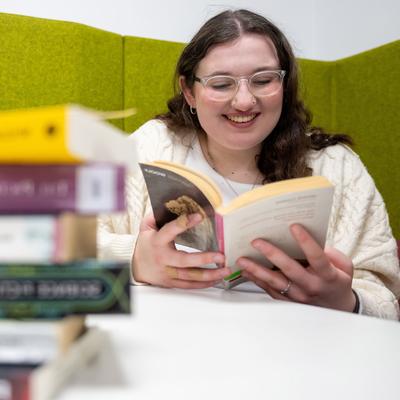 A person with glasses and long hair is smiling while reading a book at a white table. A stack of various books is in the foreground. The background features green padded walls.