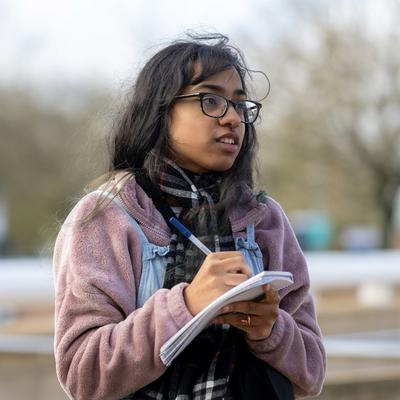A person with long hair and glasses is outdoors, wearing a pink jacket and holding a notepad and pen. They seem to be taking notes or sketching. The background is blurred with trees and a structure visible.