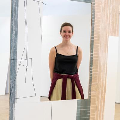 A woman stands smiling amidst an abstract art installation with geometric designs on large panels. She is wearing a black top and has a sweater tied around her waist. The setting appears to be an art gallery.