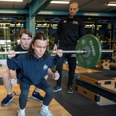 A person is performing a squat with a barbell in a gym. Another person behind them is spotting to assist, while a third person observes the exercise. The gym has weightlifting equipment and wooden platforms.