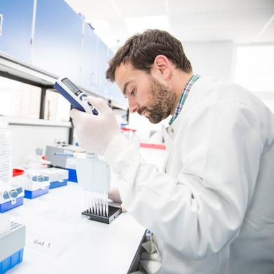 A scientist in a lab coat uses a pipette to transfer liquid into a tray, surrounded by lab equipment and bottles.