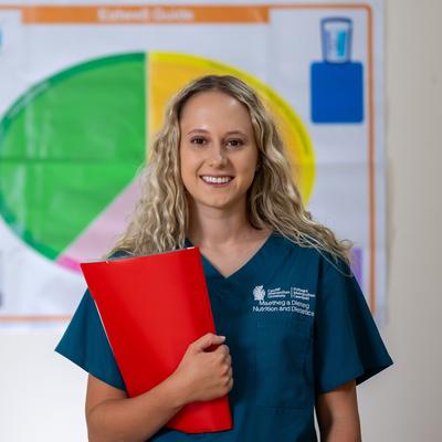 A woman in a teal uniform smiles while holding a red folder. She stands in front of a colourful chart, likely related to nutrition or dietetics. Her long blonde hair is loose, and she appears confident and professional.
