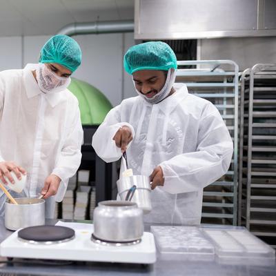 Two people in protective clothing and hairnets work in an industrial kitchen. One person holds a pot while the other pours ingredients into it. Stainless steel racks, equipment, and containers surround them.