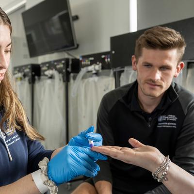 A woman in a blue hoodie and blue gloves is examining the hand of another person. A man in a black shirt observes the process. They are in a room with medical equipment visible in the background.
