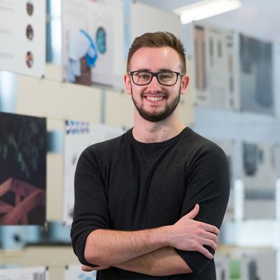 A man wearing glasses and a black sweater stands smiling with his arms crossed in front of display boards featuring various design projects and abstract images. The background is a well-lit room with natural light.