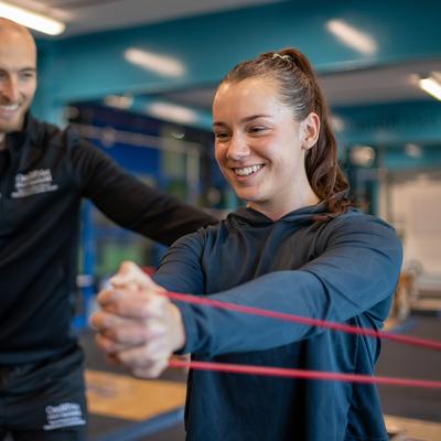 A woman exercises with a red resistance band, held in both hands, as a man in a black tracksuit smiles and assists her. They are in a gym with blue and silver equipment in the background.