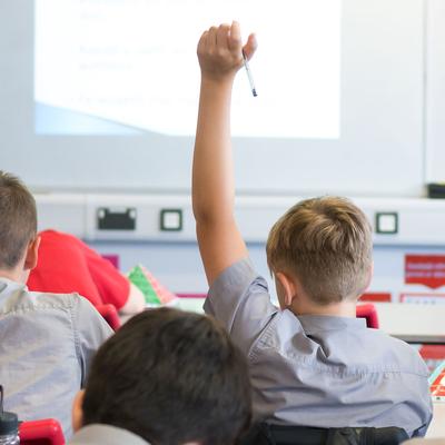 Two pupils are sitting in a classroom. One pupil has his hand raised.