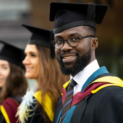 A graduate in cap and gown. In the background are three other graduates also wearing caps and gowns.