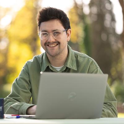 A young man sitting outside at a table. In front of him is a laptop computer and coffee mug.