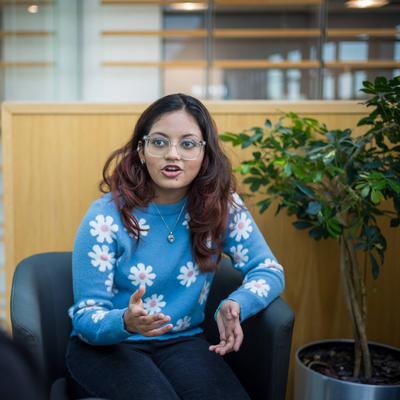 A young woman with glasses and long hair, wearing a blue sweater with white flower patterns, gestures while talking. She is seated in a modern indoor space, facing another person whose back is to the camera. A green plant is in the background.