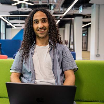 A young man sits on a green seating area with a laptop computer on his lap. In the background are rows of computer workstations.