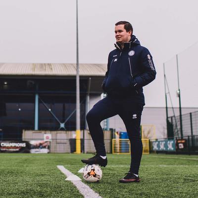 A football coach standing on the edge of the goal area with his foot on a ball.