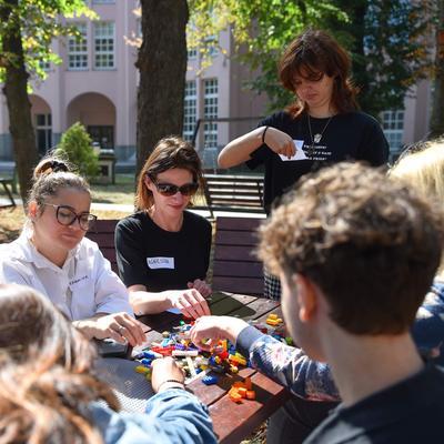 A group of people wearing name badges sit around a table outdoors. On the table in front of them are LEGO bricks.